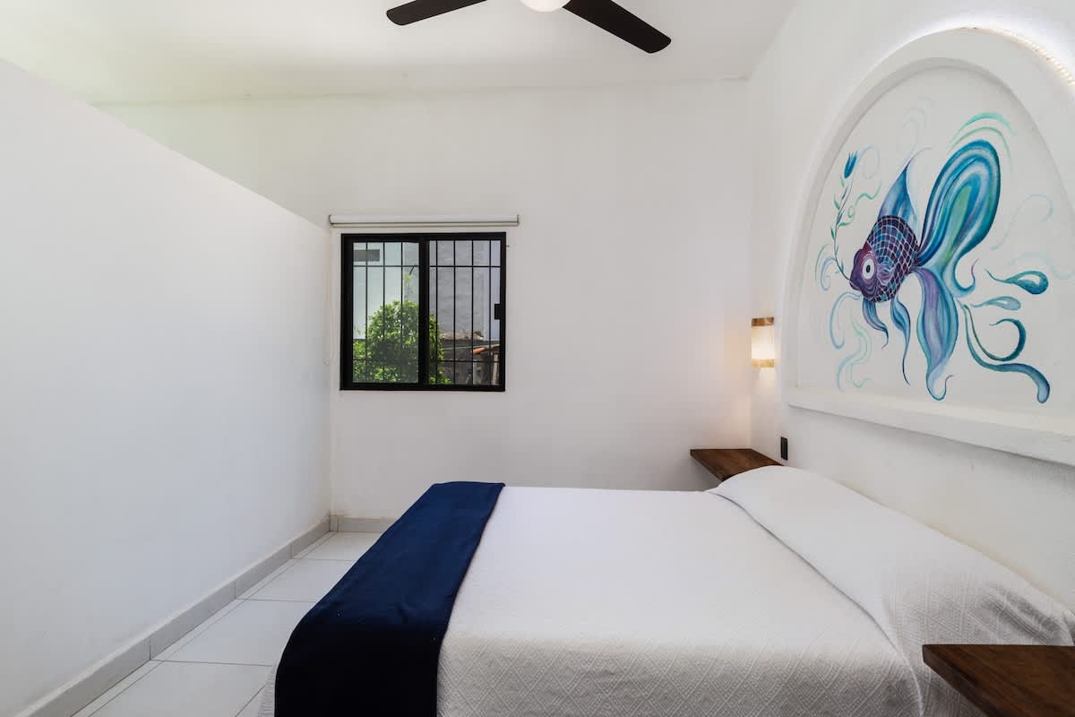 A serene bedroom with white walls, a queen bed adorned with a white comforter and blue throw, and a distinctive arched headboard featuring vibrant blue fish artwork. A window with grates provides natural light.
