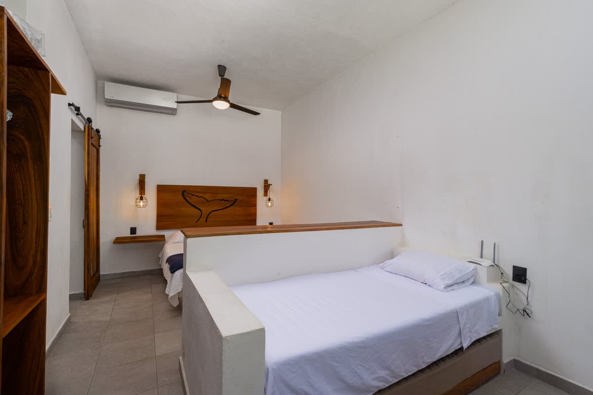 A sleeping nook with a single bed and white bedding, separated by a low partition wall. In the background, a queen bed with a distinctive wooden whale-tail headboard is visible.