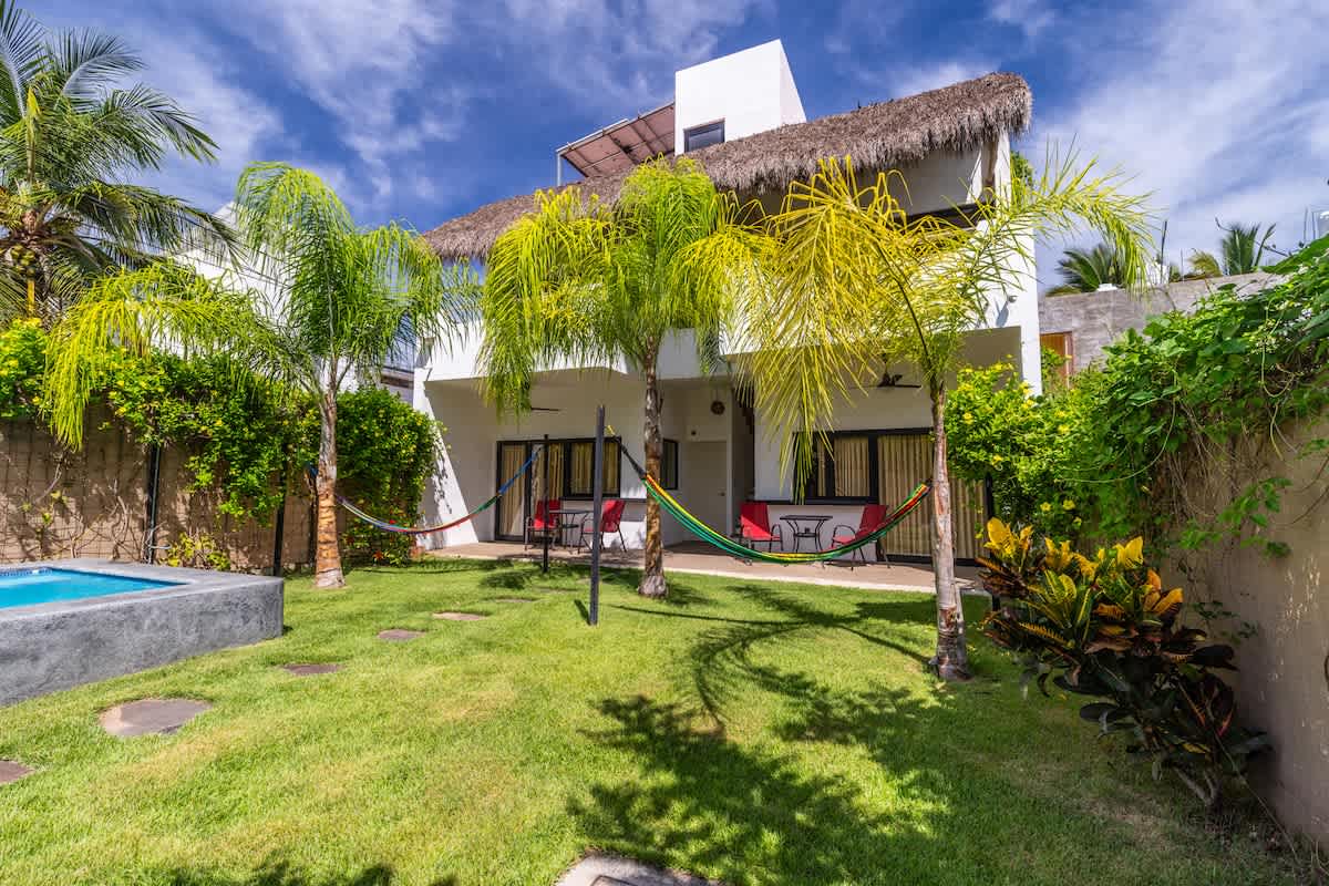 A view of the backyard showing a well-maintained green lawn with stepping stones, framed by palm trees and vibrant green foliage. The white two-story house with a thatched roof and hammocks is visible in the background.
