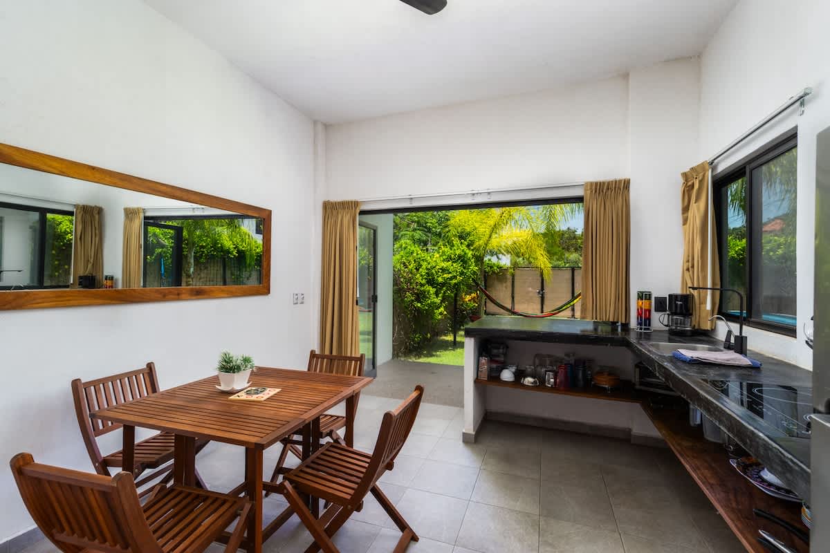 A bright dining area with a wooden square table and four matching chairs, situated next to a compact kitchen counter. Large sliding glass doors open to a lush green backyard.