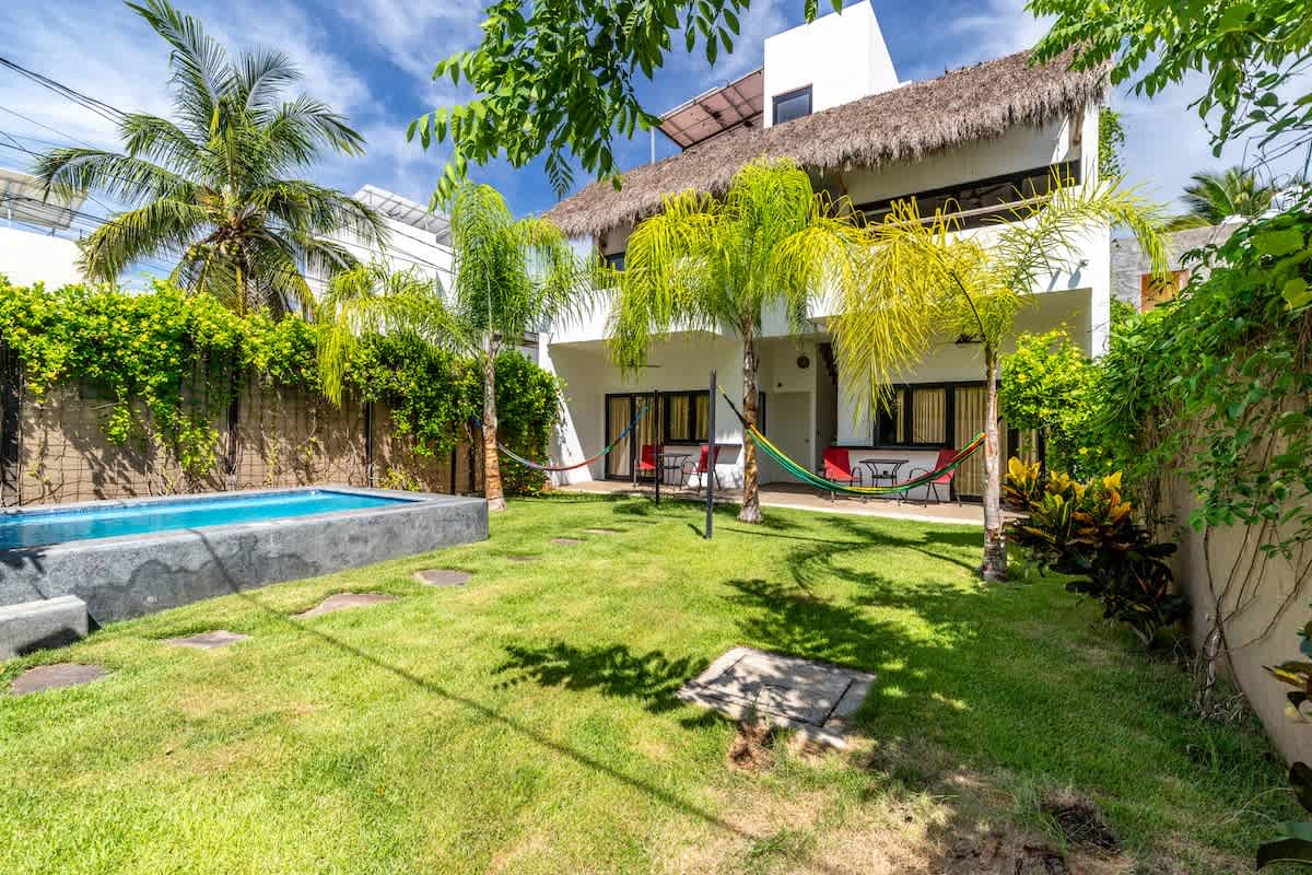 A vibrant green lawn with stepping stones leads to the back of a white two-story house featuring a thatched roof and multiple hammocks strung between palm trees. A rectangular swimming pool with blue tiles and a concrete edge is visible on the left.