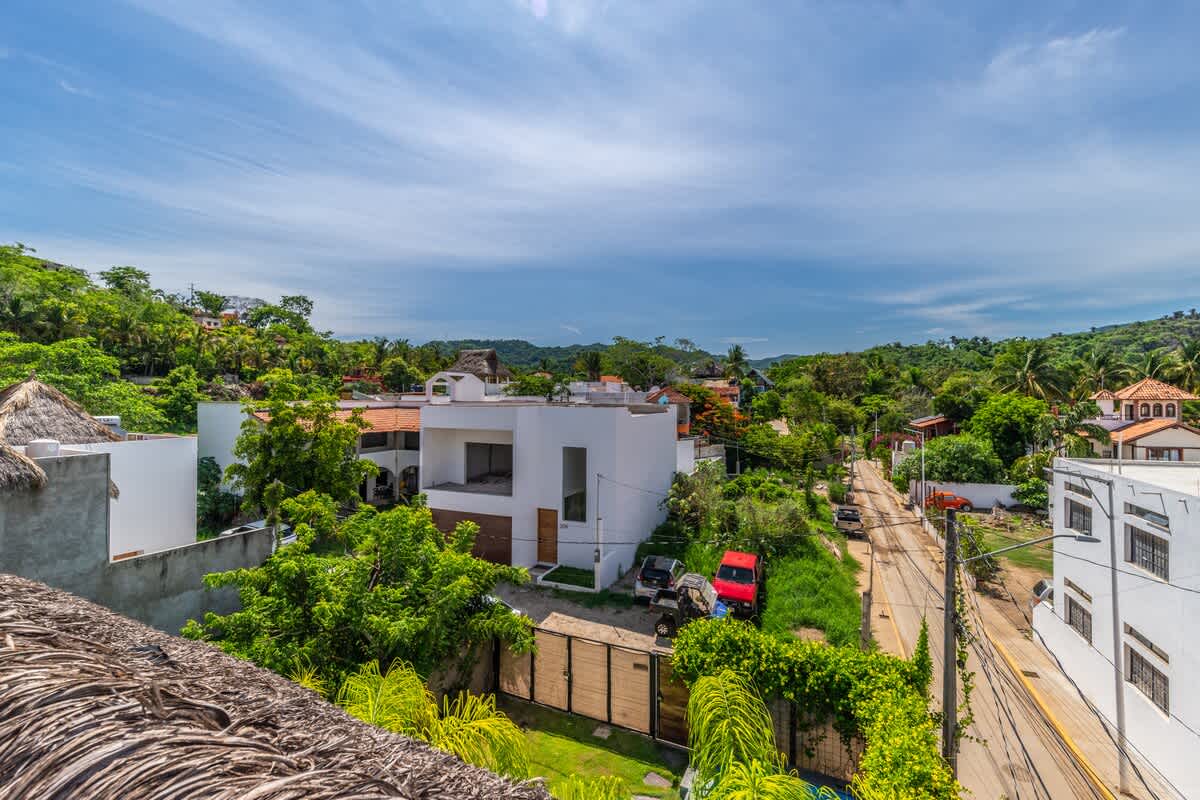 Panoramic elevated view showcasing a tropical residential area with numerous white and colorful houses nestled among dense green foliage and palm trees. A road winds through the landscape under a bright blue sky.