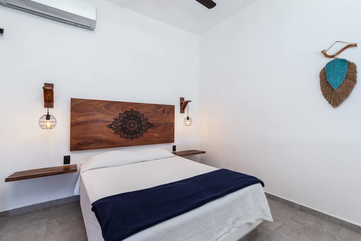 A bright bedroom features a large bed with a dark blue throw blanket and a distinctive wooden headboard with an intricate carved design, flanked by floating nightstands and industrial-style wall sconces, creating a serene retreat.