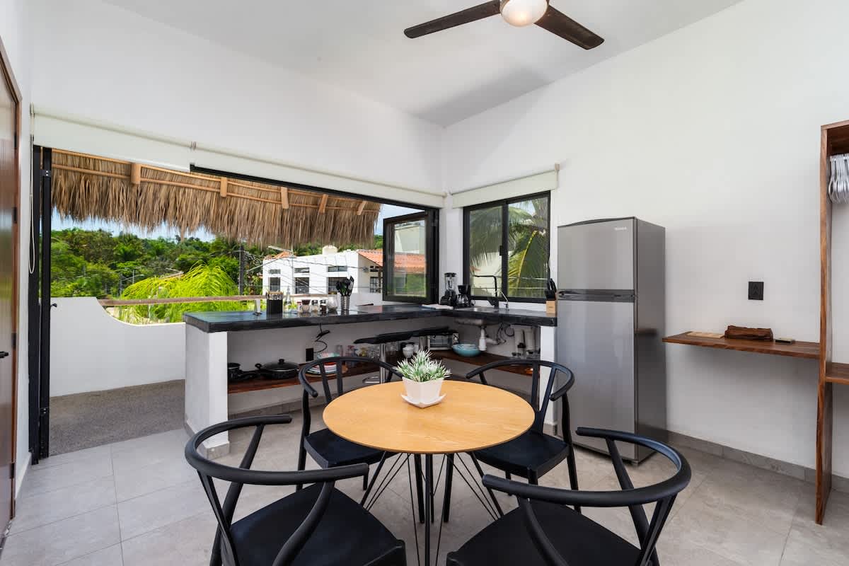 Spacious indoor-outdoor kitchen and dining area featuring dark concrete countertops, a stainless steel refrigerator, and a round wooden dining table with black chairs. Large windows and an open wall connect to the tropical exterior.
