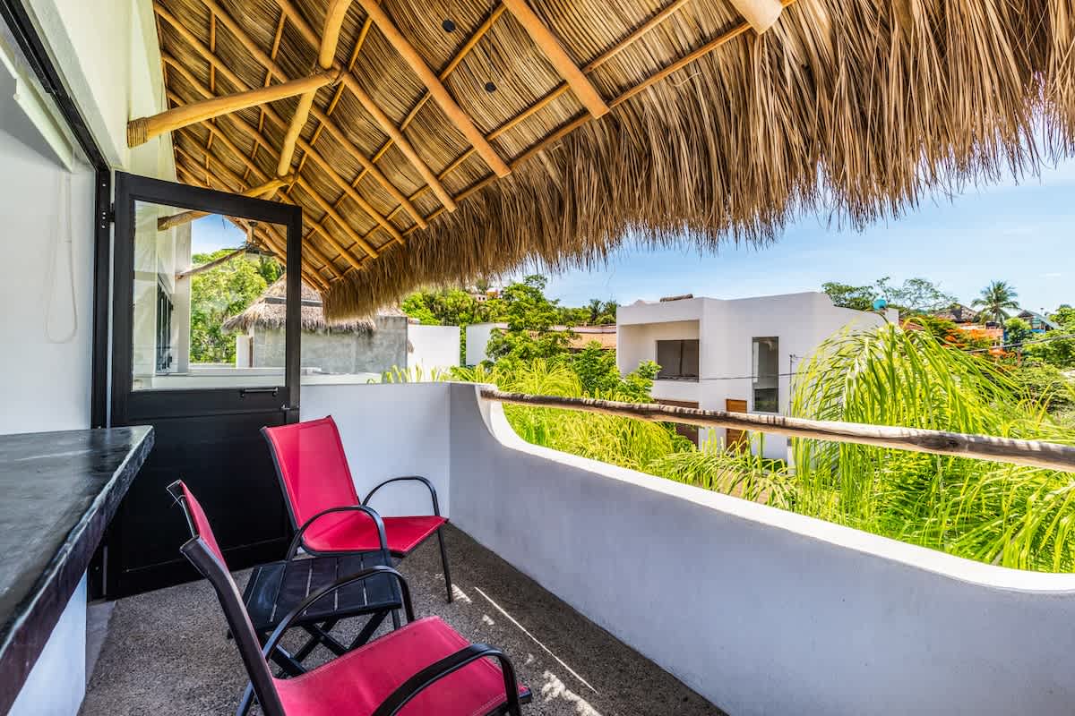 Inviting balcony with a traditional thatched roof (palapa) providing shade over a dark countertop bar and red outdoor chairs. The railing offers views of the surrounding tropical houses and lush greenery.