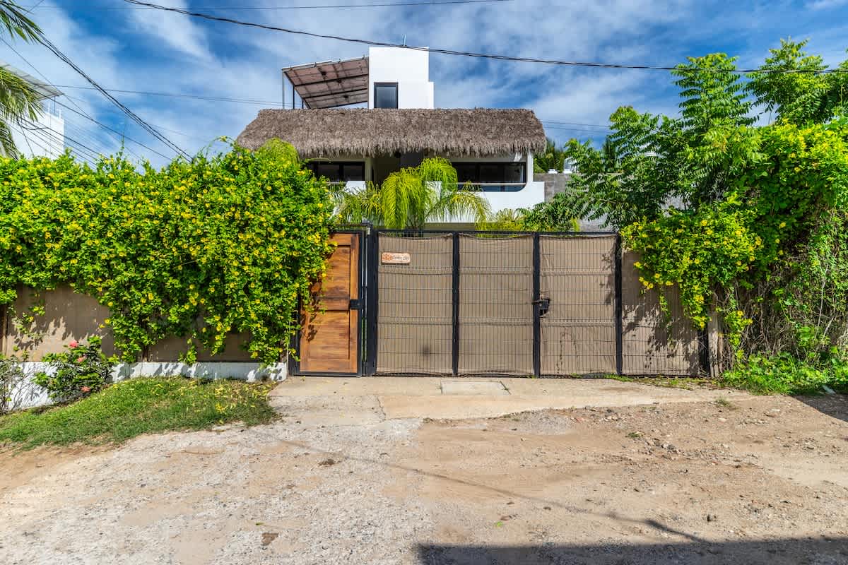 The front of a white two-story house with a thatched roof, partially obscured by lush green climbing plants on a concrete wall and a dark metal gate. A wooden pedestrian gate is integrated into the metal gate, and a dirt and gravel driveway leads up to it.