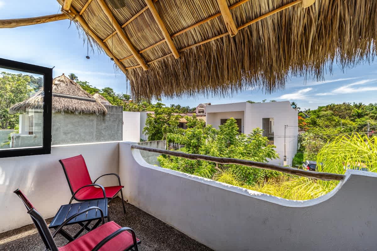A scenic view from a balcony with a rustic thatched roof, showing lush green trees and other white buildings under a bright blue sky. Two red chairs and a small table are visible on the balcony.