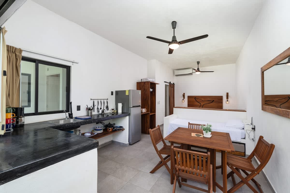 An open-plan studio space showcasing a compact kitchen with dark countertops, a stainless steel refrigerator, and a wooden dining table with four chairs. A queen bed is visible in the background.