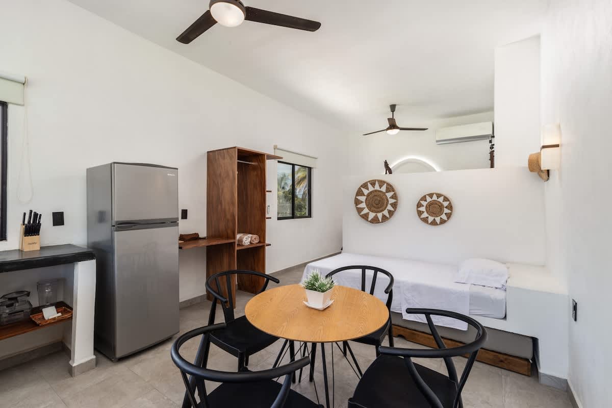 A bright studio apartment with a single bed, a round wooden dining table with four black chairs, and a compact kitchen area featuring a stainless steel refrigerator and a wooden storage unit. Decorative woven plates adorn the wall.