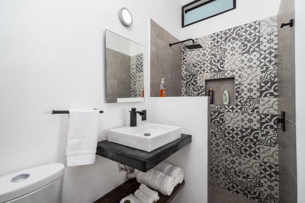 A modern bathroom with a white vessel sink on a dark floating vanity, a toilet, and a shower area featuring patterned grey and white tiles and a black rainfall showerhead. White towels are neatly stacked below the sink.