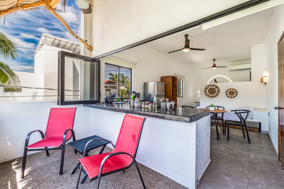 Open-air balcony featuring a dark countertop bar with red outdoor chairs, overlooking a lush tropical landscape. The space seamlessly connects to an indoor area with a small dining table and decorative wall art.