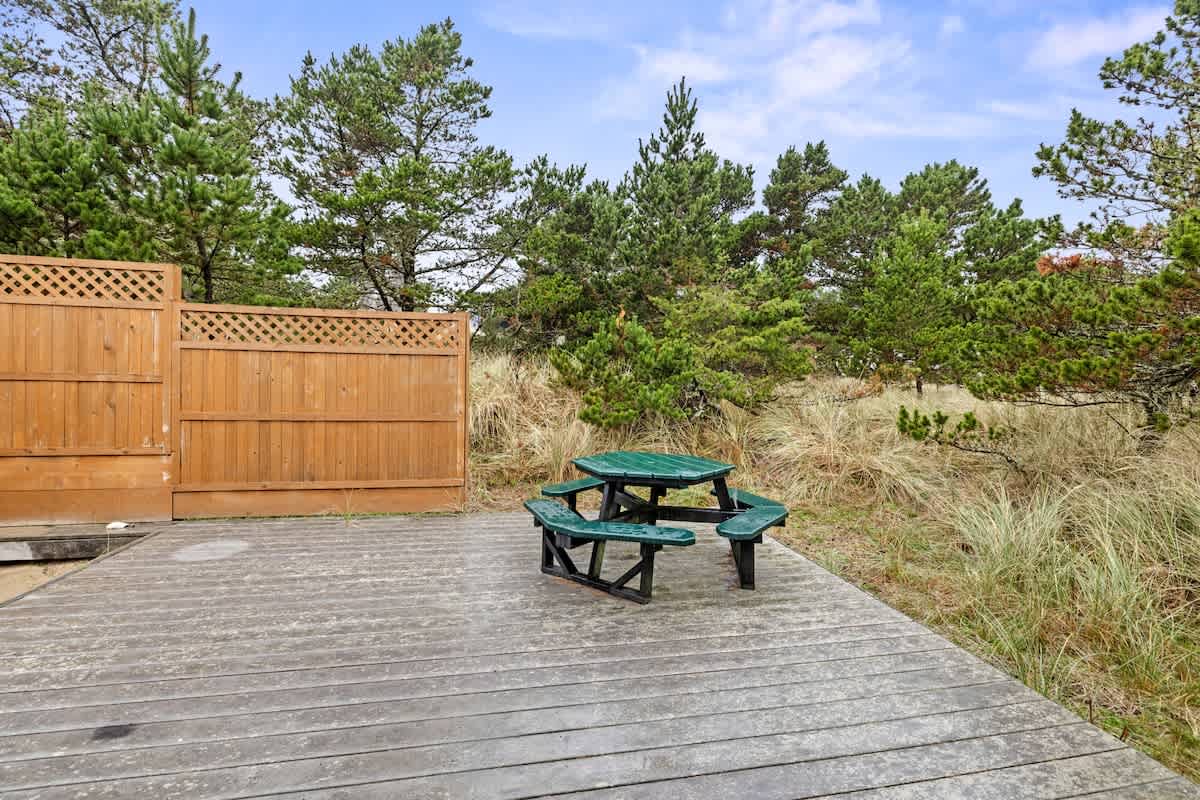 An expansive weathered wooden deck extends into a natural landscape with tall grasses and pine trees. A green octagonal picnic table with attached benches sits on the deck, offering an outdoor dining spot.