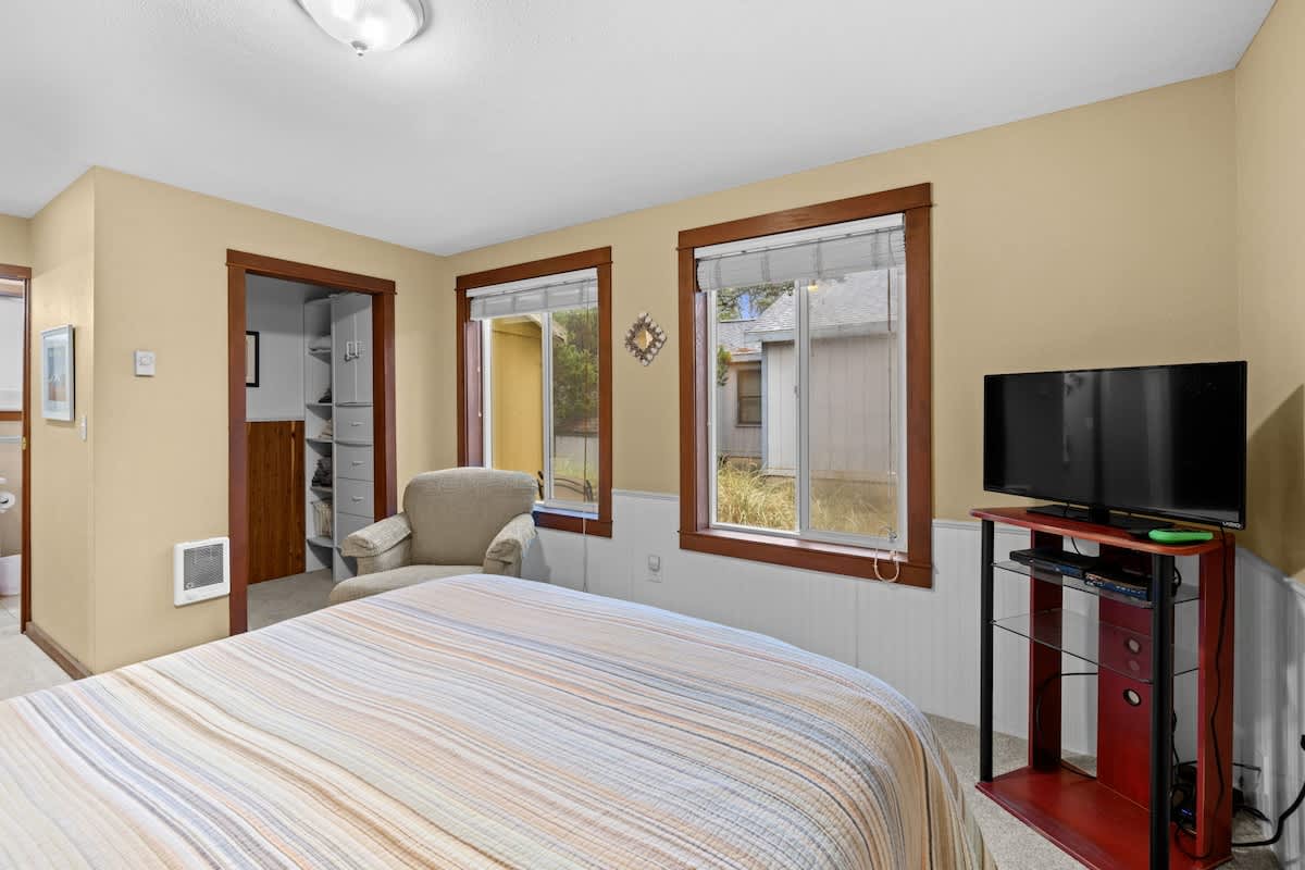 Another perspective of bedroom-3, featuring the bed with a striped comforter, an armchair, and two windows. A doorway with dark wooden trim leads to an adjacent room, and the light yellow walls with wainscoting are visible.