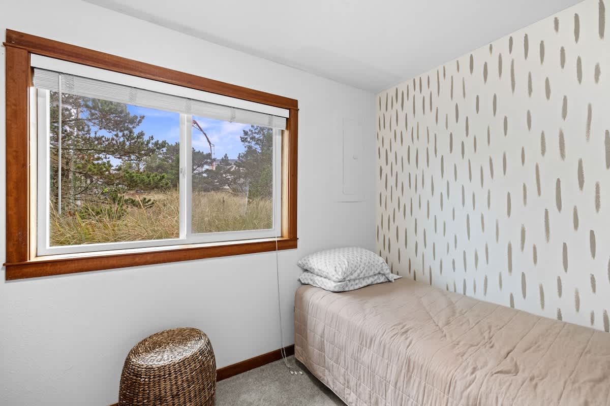 A cozy bedroom featuring a single bed with a light brown quilt, white walls, and one accent wall with a subtle dotted pattern. A large window with warm wooden trim overlooks a natural, grassy landscape.