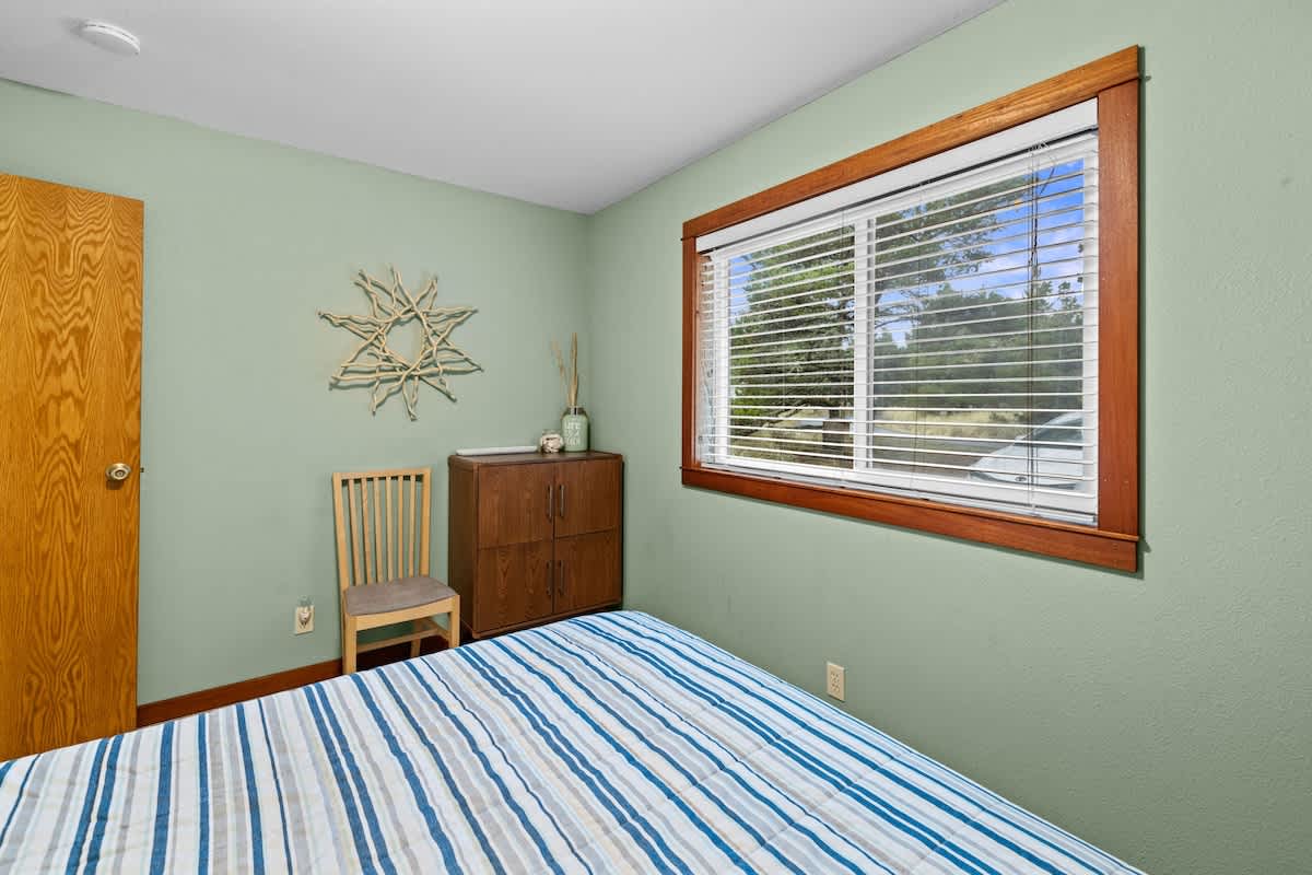Another view of bedroom-2, highlighting the light green walls, the bed with a blue striped comforter, and a wooden dresser with decorative items. A window with wooden trim provides natural light.