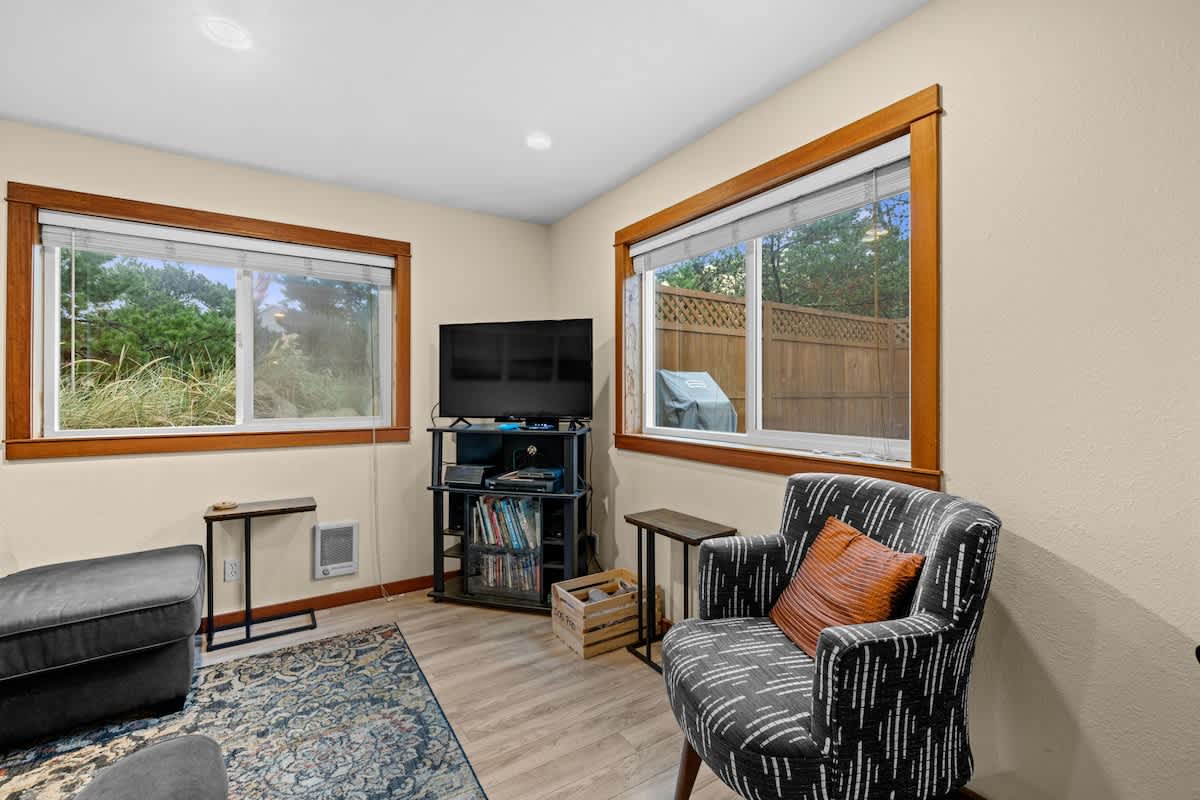 Cozy living room with light beige walls, wood-look laminate flooring, and two large windows with natural wood trim. A black television is on a stand, flanked by a dark patterned armchair and a dark ottoman.