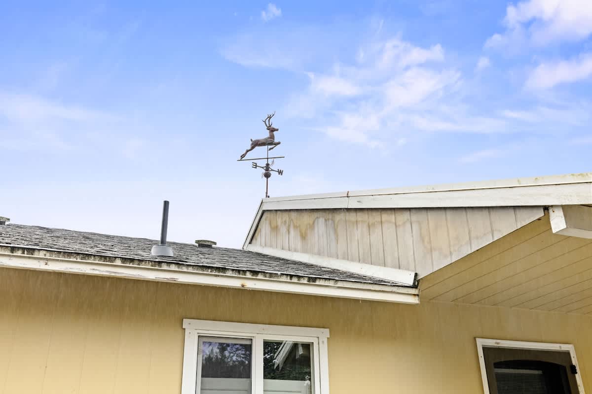 A close-up view of the house's roofline, featuring a dark shingle roof and light yellow siding. A decorative deer weathervane with cardinal directions is prominently displayed against a bright blue sky with scattered clouds.