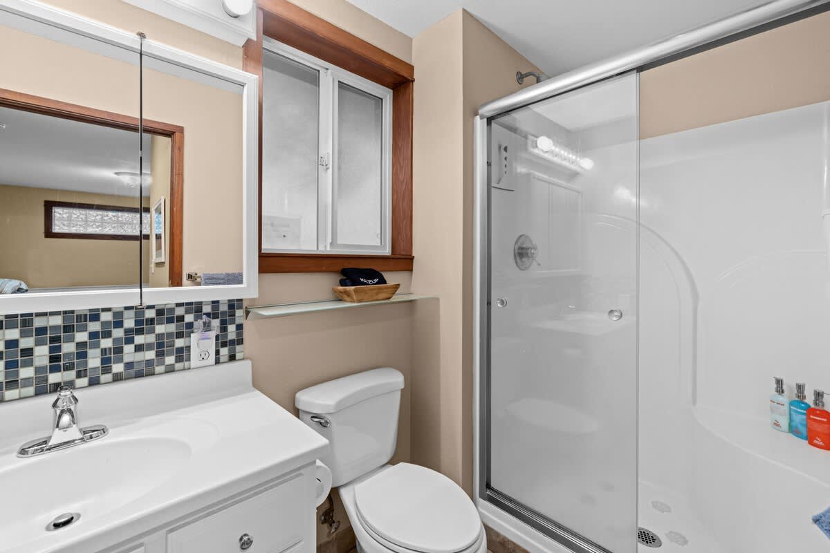 Full view of bathroom-1, showcasing a white vanity with a sink, a mosaic tile backsplash, a toilet, and a glass-enclosed shower. A window with wooden trim is above the toilet.