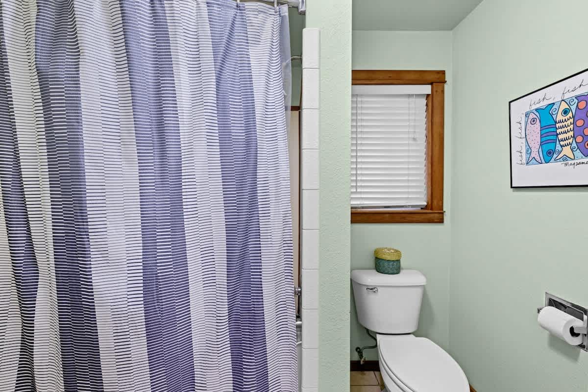 Close-up of a bathroom area featuring a white toilet, a window with white blinds, and a blue and white striped shower curtain. The walls are painted a light mint green.