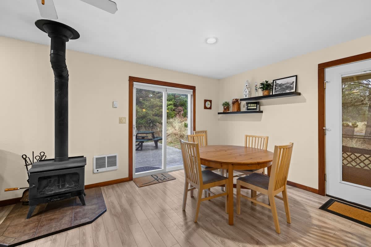 A cozy dining area features a round wooden table with four chairs, positioned near a black wood-burning stove with a stovepipe extending upwards. A sliding glass door leads to an outdoor deck, and light wood flooring completes the space.