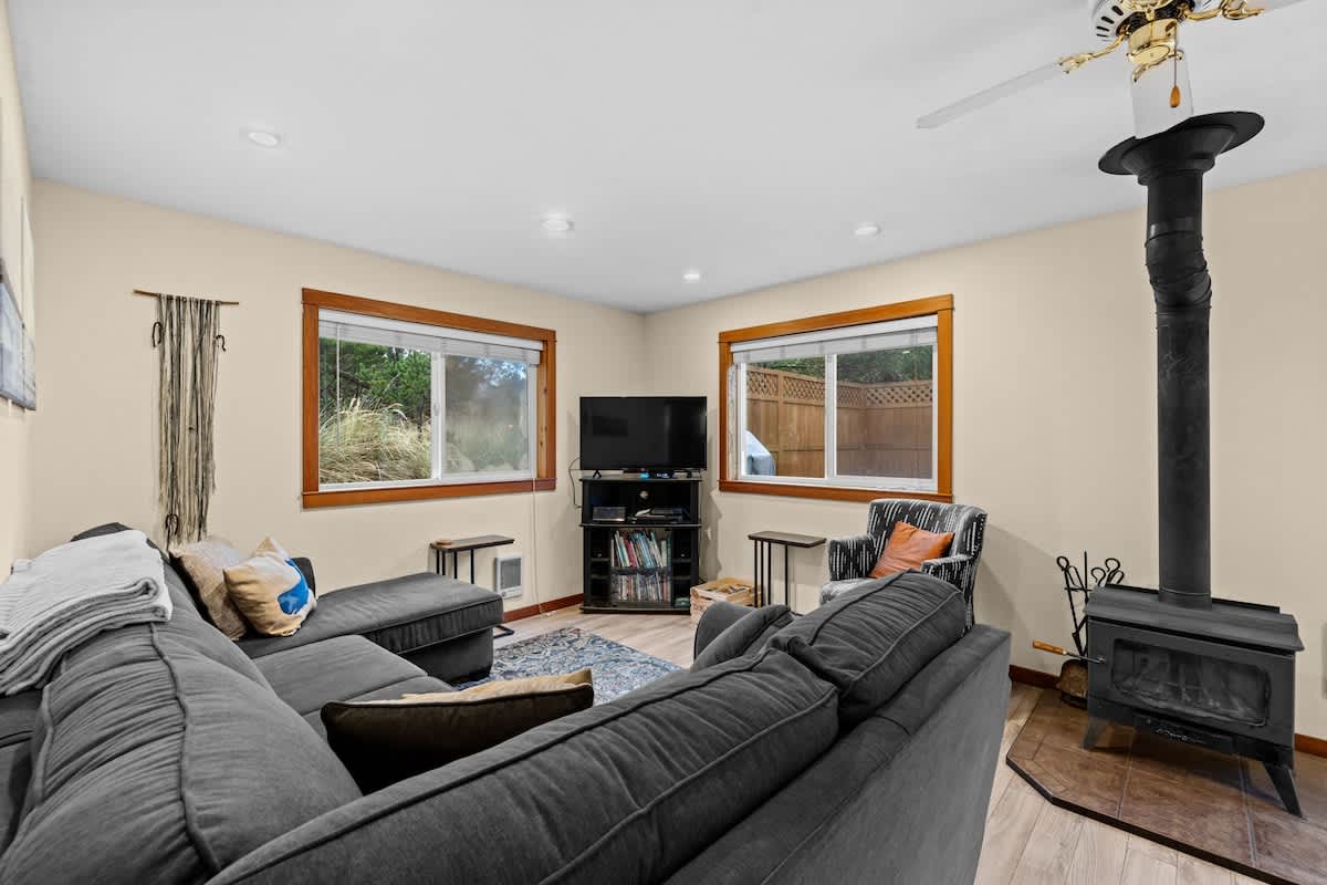 Spacious living room with light beige walls, wood-look laminate flooring, and a large dark gray sectional sofa. A black wood-burning stove with a tall pipe stands in the corner, next to a ceiling fan.
