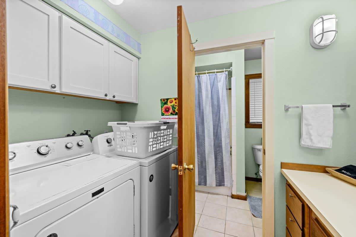Laundry area featuring a white stacked washer and dryer unit beneath white upper cabinets, set against light mint green walls. It opens into a bathroom with a toilet and shower.