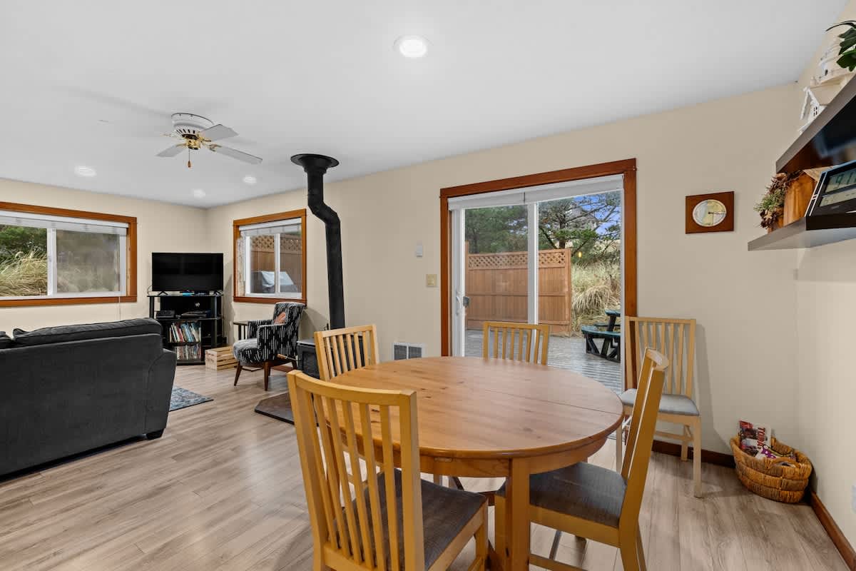 An inviting open-concept living area features a dark gray sectional sofa, a black wood-burning stove, and a round wooden dining table with four chairs. Light wood flooring extends throughout the space, with large windows offering views of the outdoors.