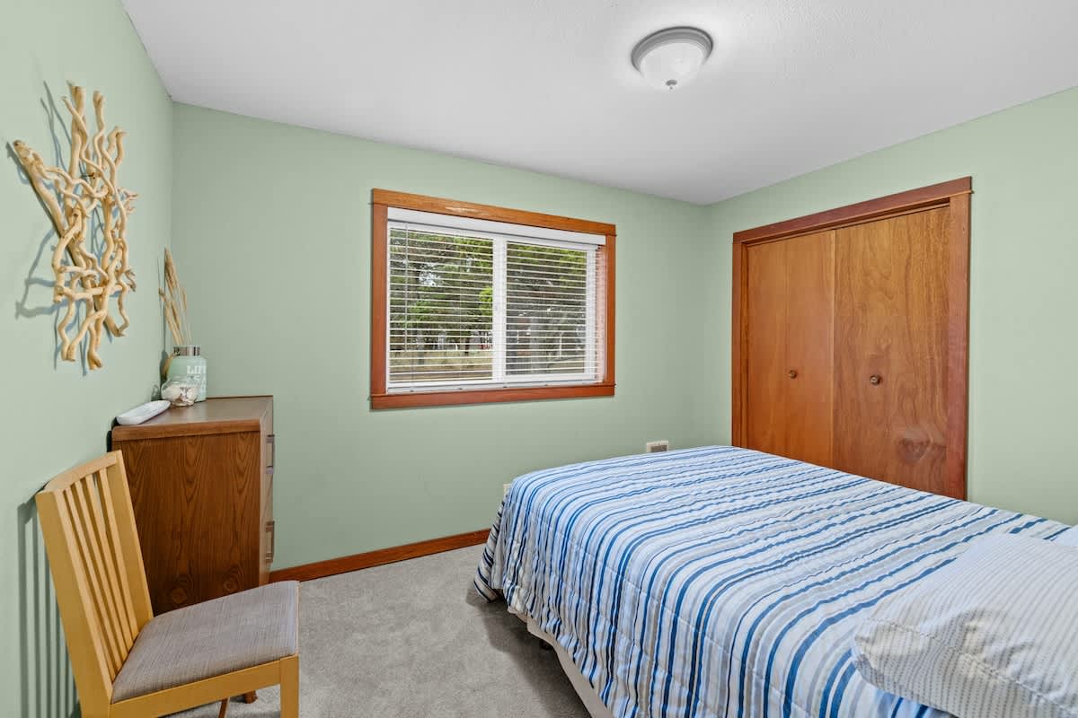 A serene bedroom with light green walls, featuring a bed with a blue and white striped comforter. A large window with wooden trim offers a view of trees, and a wooden closet is visible.