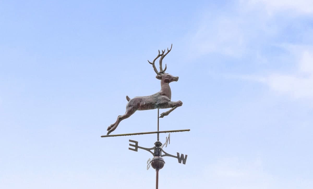 A detailed close-up of a bronze-colored weathervane shaped like a leaping deer with antlers, mounted atop a directional arrow indicating East, West, and South. It stands out against a clear, bright blue sky.