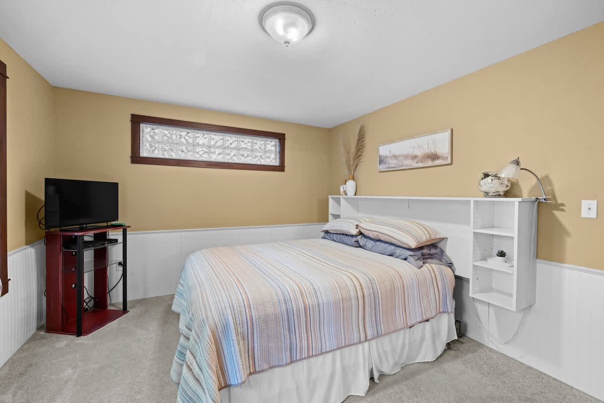 View of bedroom-3 showcasing the bed with a multi-colored striped comforter and a white headboard with integrated shelving. The light yellow walls and white wainscoting create a cozy atmosphere, with a TV on a red stand.