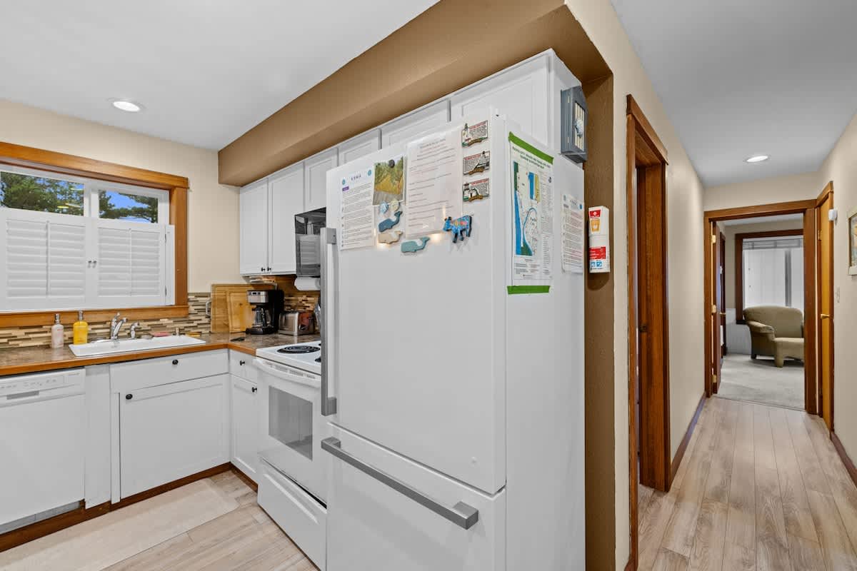 Bright kitchen with white cabinets and appliances, including a refrigerator and stove, set against light beige walls. Wood-look laminate flooring extends into a hallway leading to other rooms.