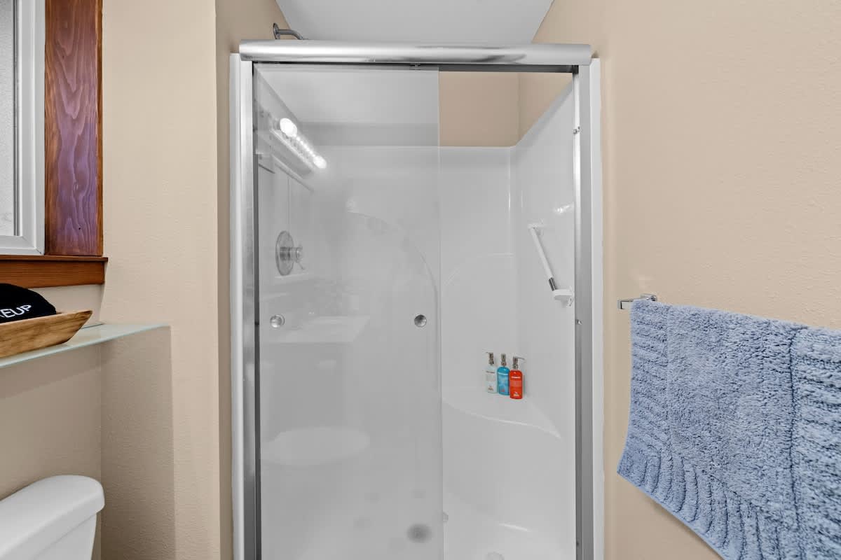 Close-up view of the shower in bathroom-1, featuring a clear glass enclosure and white shower walls. Toiletries are visible inside the shower, and a blue towel hangs nearby.