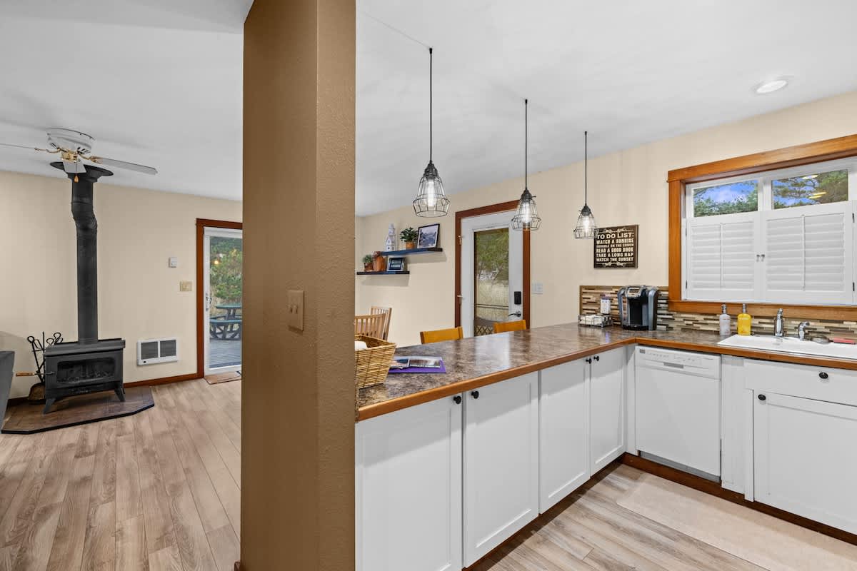 Open concept kitchen with a long counter and industrial-style pendant lights, connecting to the living area. White cabinets and wood-look flooring are visible, with a glimpse of the wood-burning stove in the adjacent room.