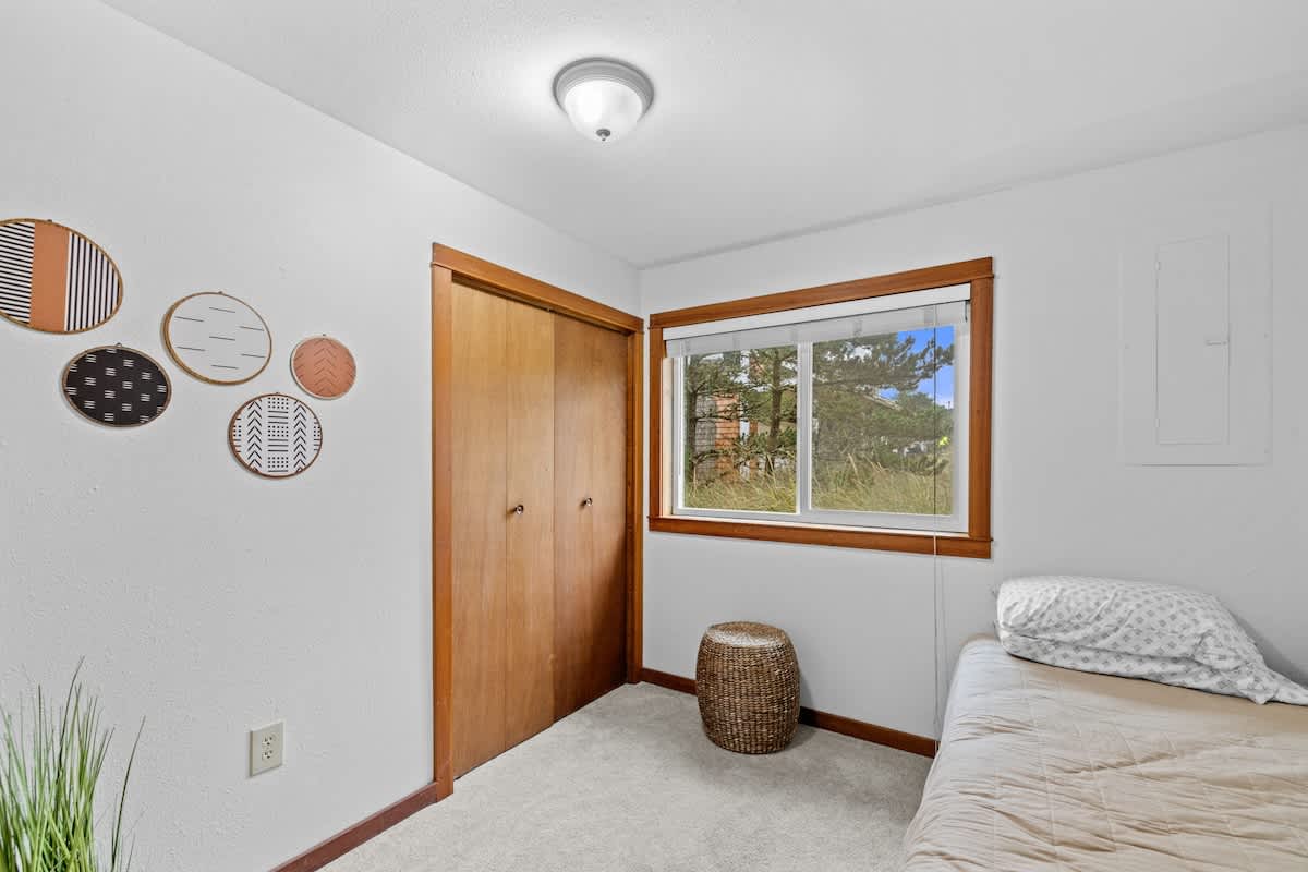 Simple bedroom with a single bed, light gray carpet, and white walls. A large wooden closet and a window with natural wood trim are prominent, along with decorative circular wall hangings.