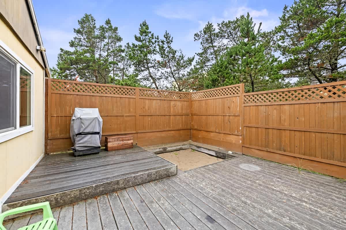 A private outdoor deck area features weathered wooden planks, a lattice-topped wooden fence, and a covered grill. A square sand pit is recessed into the deck, surrounded by tall pine trees under a blue sky.