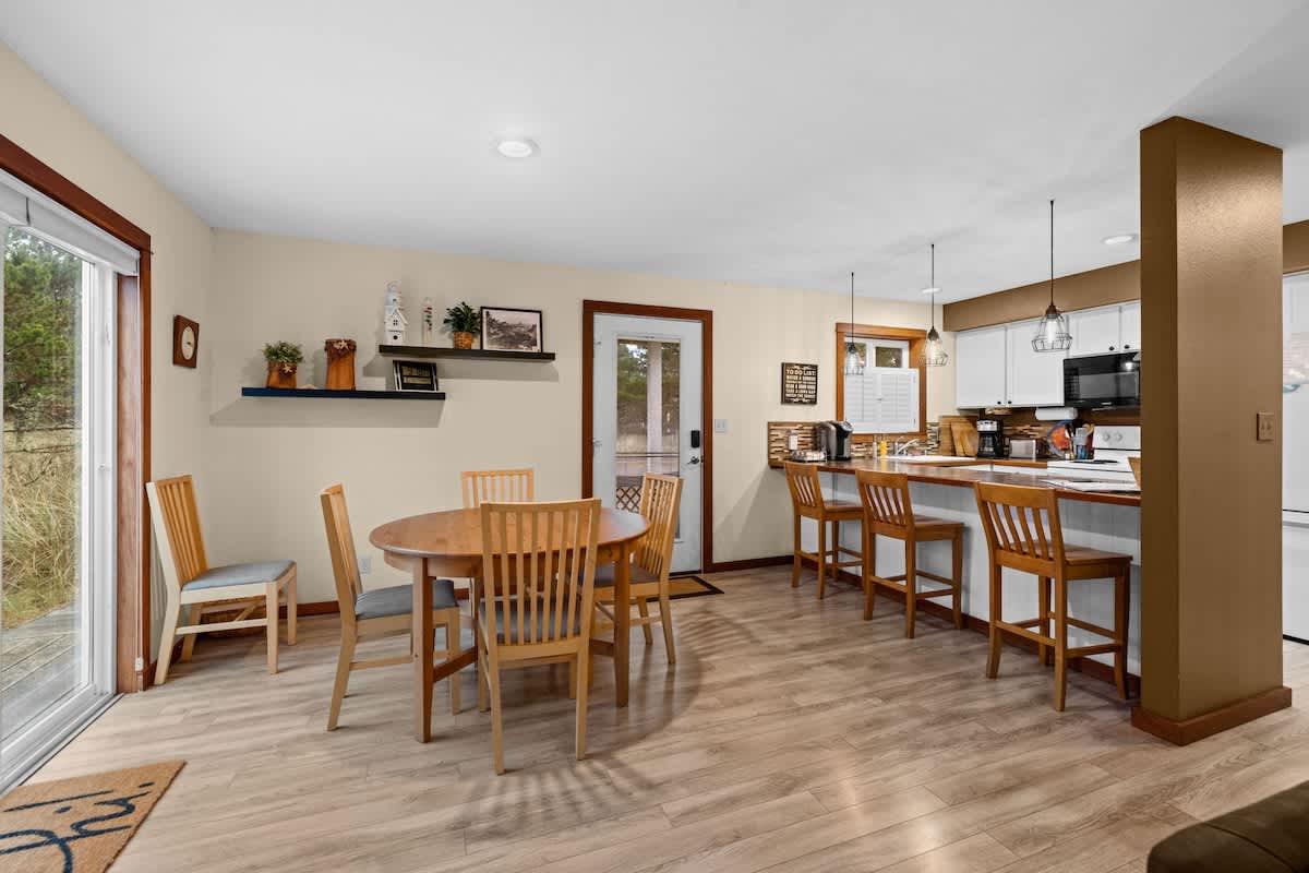 An open-concept dining area features a round wooden table with four chairs, flowing into a modern kitchen with a breakfast bar. Light wood flooring and cream walls create a bright atmosphere, with a glass door leading to an outdoor deck.