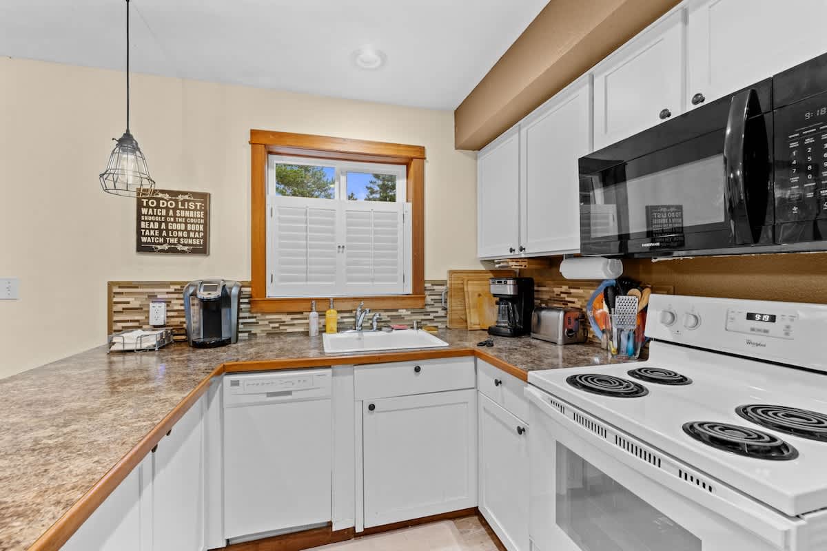 Kitchen counter area with white cabinets, a white sink, and white appliances including a dishwasher, stove, and microwave. A window with white shutters is above the sink, and a coffee maker is on the counter.