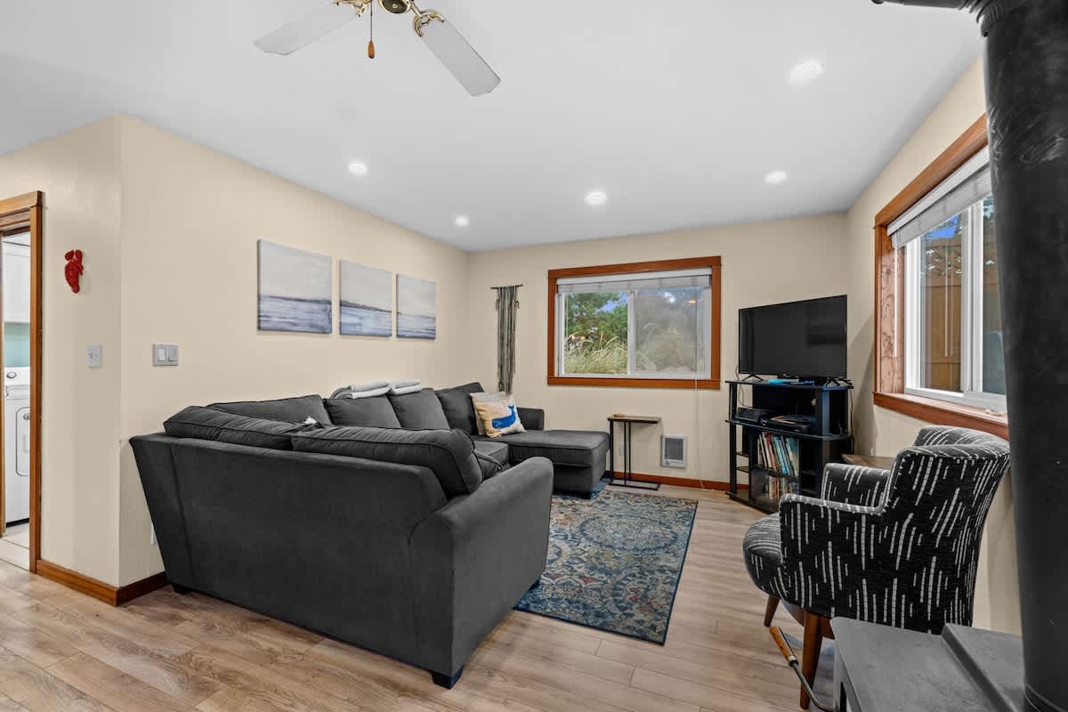 A comfortable living room features a large dark gray sectional sofa, a flat-screen TV on a stand, and a patterned accent chair. Light wood flooring is covered by a blue and white patterned rug, and large windows provide natural light.