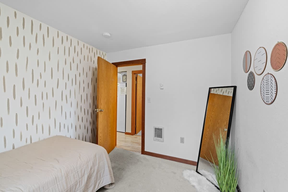 View of bedroom-1 showing the light brown quilted single bed, the patterned accent wall, and a full-length mirror leaning against a white wall. A wooden door leads to another part of the house.