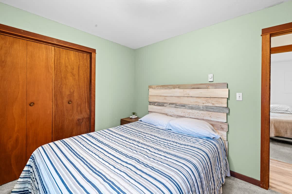 Bedroom with a double bed featuring a rustic wooden headboard, light gray carpet, and light mint green walls. A large wooden closet is visible, and an open doorway leads to another room.