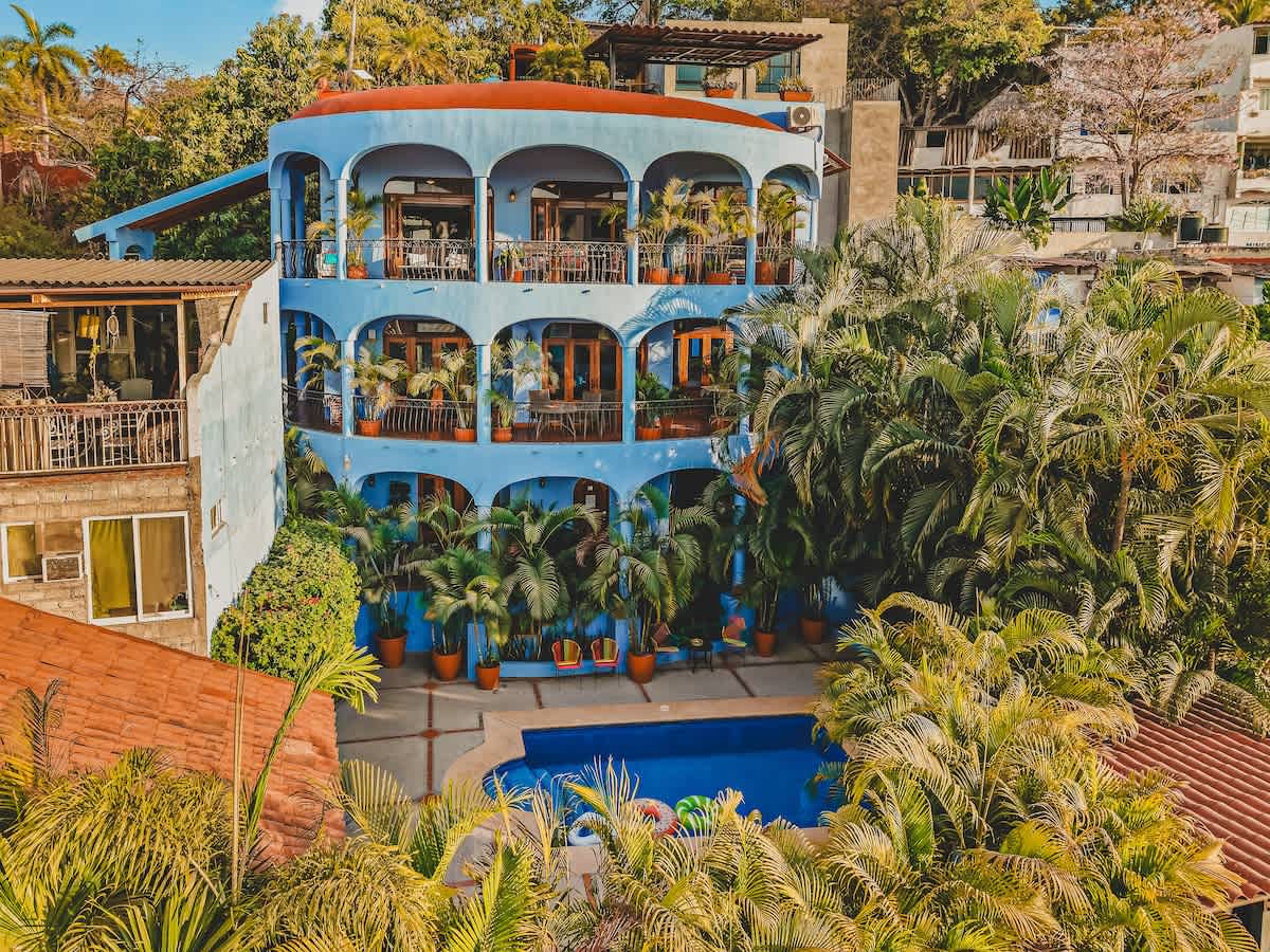 An aerial view of a large, light blue multi-story building with arched balconies, surrounded by dense tropical foliage and palm trees, with a bright blue swimming pool visible in the foreground.