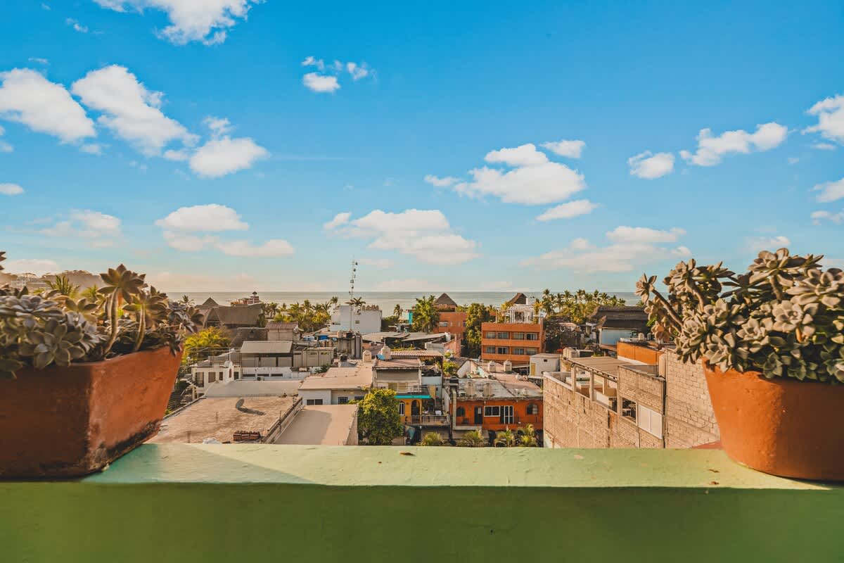 A panoramic view from a high vantage point, showcasing a colorful coastal town with various buildings and lush greenery, extending to a calm ocean under a bright blue sky with scattered clouds.