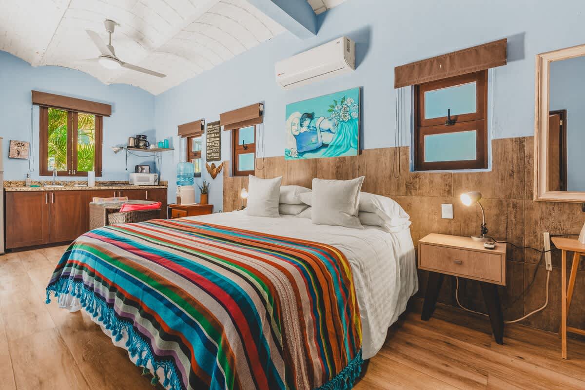 A light blue studio bedroom featuring a large bed with a colorful striped blanket, a small kitchenette with dark wood cabinets, and multiple windows with wooden frames. A ceiling fan and an AC unit are visible.