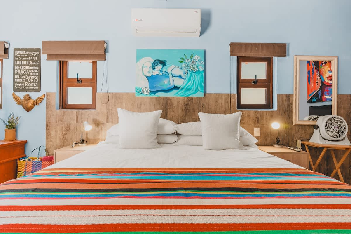 A vibrant bedroom with light blue walls and a wooden half-wall, featuring a large bed with crisp white linens and a colorful striped blanket. Two small windows with brown blinds flank a light blue artwork above the wooden headboard.