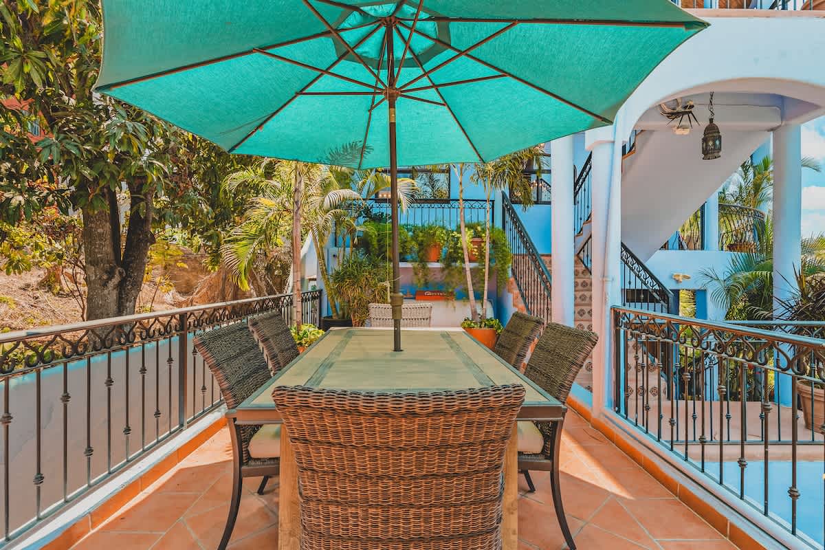 An outdoor dining area on a tiled terrace, featuring a large wicker dining table with six chairs and a turquoise umbrella, surrounded by a black metal railing and lush greenery.