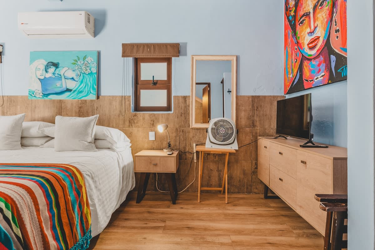 A cozy bedroom area with light blue walls and a wooden half-wall, featuring a bed with a colorful blanket and two nightstands with lamps. A small floor fan, a full-length mirror, and a dresser with a TV are visible on the right.