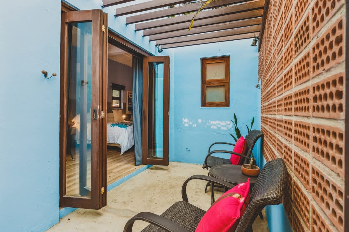 A private outdoor patio with light blue walls and a decorative brick screen, featuring two dark wicker chairs and a small table, with wooden bi-fold doors opening into a bright room with a bed visible.