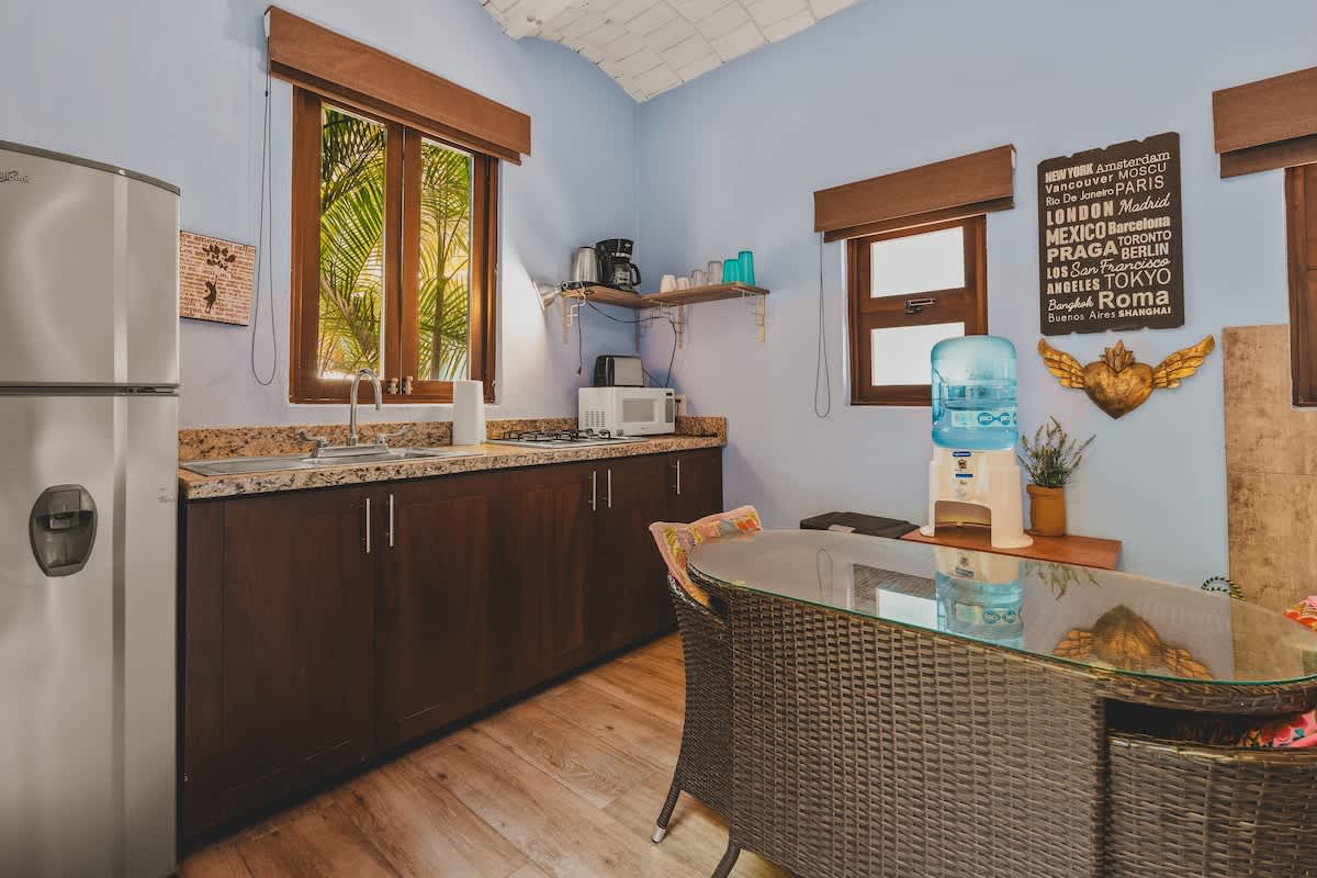 A compact kitchenette with dark wood cabinets, a granite-look countertop, a stainless steel refrigerator, microwave, coffee maker, and a water dispenser. A glass-top dining table with wicker chairs is also present.