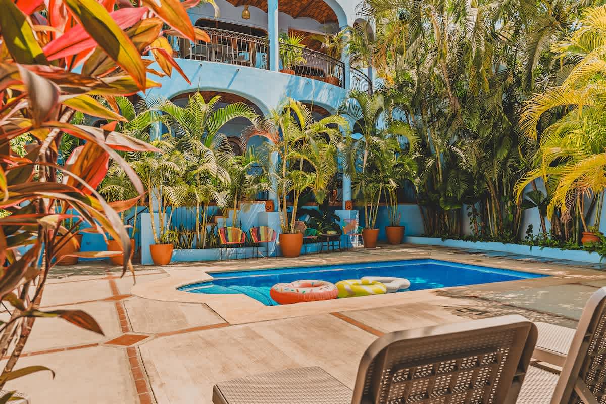 An inviting outdoor swimming pool with clear blue water and inflatable rings, set within a tiled patio area, surrounded by abundant tropical foliage and palm trees, with lounge chairs in the foreground.