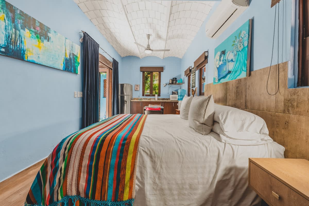 A long shot of a studio apartment, showcasing a large bed with a colorful striped blanket in the foreground and a compact kitchen area visible in the background. Light blue walls, a vaulted brick ceiling, and various artworks create a vibrant and inviting atmosphere.
